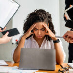 A stressed woman at a desk surrounded by multiple devices and people