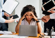 A stressed woman at a desk surrounded by multiple devices and people