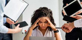 A stressed woman at a desk surrounded by multiple devices and people