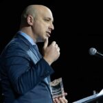 A man in a blue suit speaking at a podium while holding an award