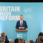 A politician delivering a speech at a podium with British flags in the background