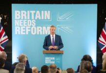 A politician delivering a speech at a podium with British flags in the background