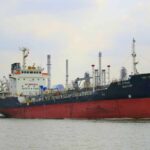 A cargo ship navigating through a port with industrial buildings in the background