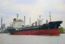 A cargo ship navigating through a port with industrial buildings in the background