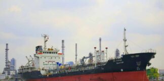 A cargo ship navigating through a port with industrial buildings in the background