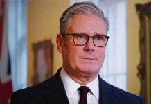 A man in formal attire with glasses, looking serious in an indoor setting