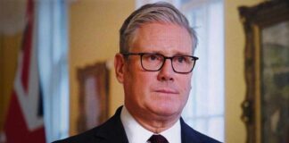 A man in formal attire with glasses, looking serious in an indoor setting