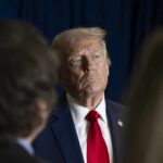 A serious-looking man in a suit with a red tie, surrounded by blurred figures