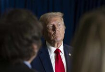 A serious-looking man in a suit with a red tie, surrounded by blurred figures