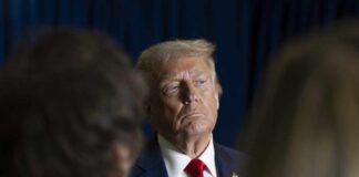 A serious-looking man in a suit with a red tie, surrounded by blurred figures