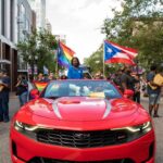 Participants in a pride parade with flags and a red convertible car