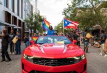 Participants in a pride parade with flags and a red convertible car