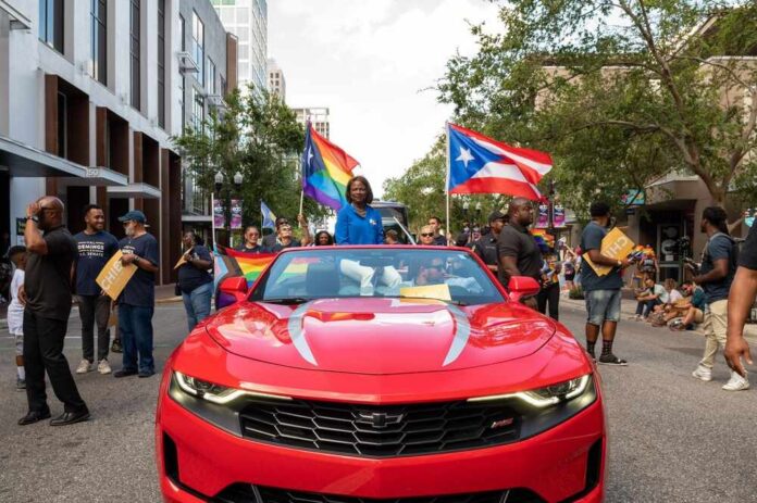 Participants in a pride parade with flags and a red convertible car