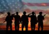 Silhouetted soldiers saluting against an American flag backdrop at sunset