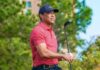 Woods’ Mysterious Treatment Journey Begins A golfer holding a club outdoors, wearing a red striped shirt