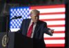 A man in a suit gestures while speaking at a podium with an American flag in the background