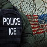 Rising ICE Violence — Are Law Enforcers Safe Anymore? Police officer in tactical gear standing near a barbed wire fence with an American flag in the background