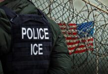 Police officer in tactical gear standing near a barbed wire fence with an American flag in the background