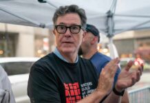 A man in a black t-shirt with a writer's guild logo, clapping at a public event