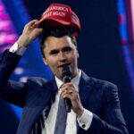 Man in a suit holding a red cap while speaking into a microphone