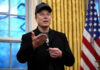 One Trillion Dollars, Countless Unanswered Questions Man wearing hat gesturing in room with flag