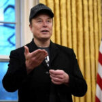 Man wearing hat gesturing in room with flag