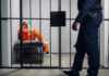 From Internet Fame To Courtroom Reality Inmate in orange uniform sitting on a jail bunk with guard walking past