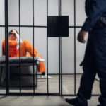 From Internet Fame To Courtroom Reality Inmate in orange uniform sitting on a jail bunk with guard walking past