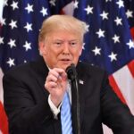 Trump Takes Immigration Fight To America’s Banks man speaking at a podium with American flags behind him