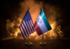 War Clips Exposed—Who’s Playing Us? Flags of the United States and Iran displayed on stands with a smoky background