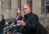 man in black suit speaking at outdoor press conference