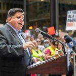 Speaker at a podium addressing a crowd with microphones and protest signs