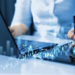 Businessperson analyzing stock data on tablet beside laptop