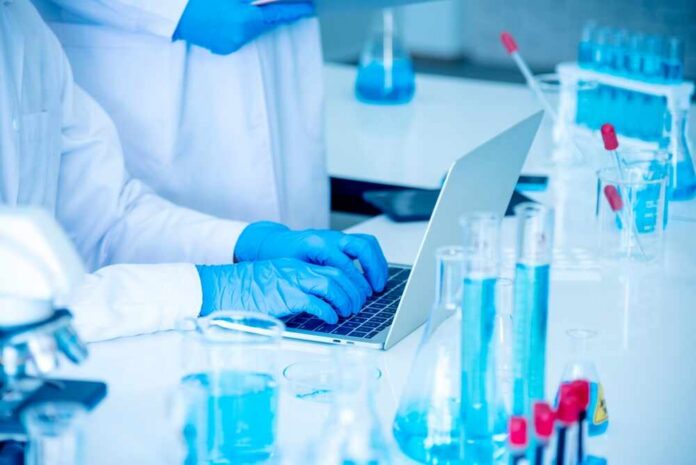 A scientist typing on a laptop in a laboratory filled with glassware