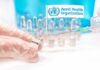 Gloved hands holding vaccine vial and syringe near WHO banner