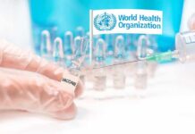Gloved hands holding vaccine vial and syringe near WHO banner