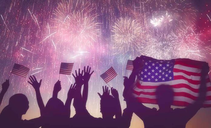 silhouetted crowd waving US flags under colorful fireworks
