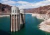 A large dam structure with two towers over a blue water reservoir surrounded by rocky mountains