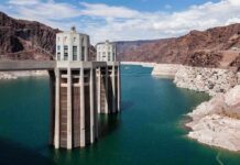 A large dam structure with two towers over a blue water reservoir surrounded by rocky mountains