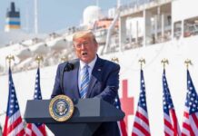 man in suit speaking at a podium with flags and a ship behind