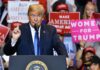 Donald Trump speaking at a rally podium with crowd holding signs