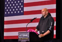 Fetterman Flip Tease Shocks Washington Man in hoodie speaking at a podium before a large U.S. flag