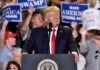 Donald Trump speaking at a rally podium with crowd holding signs behind him