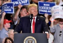 Donald Trump speaking at a rally podium with crowd holding signs behind him